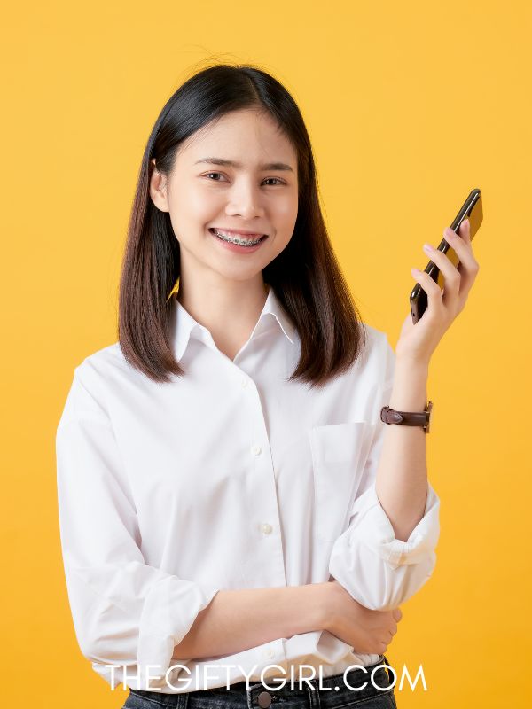 A yellow background with a teen girl wearing a white shirt holding a phone in her hand. She has medium length brown hair and braces and is smiling at the camera. 