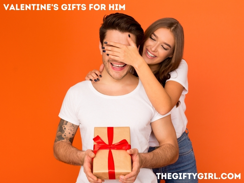 Orange backdrop with a man sitting down with a present in his hands. A woman is standing behind him wit her hand over his eyes. They are both happy. Text overlay says "Valentine's Gifts for Him TheGiftyGirl.com"