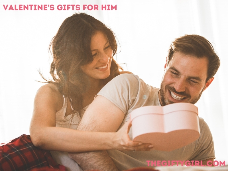 Man lying on his side in a bed being presented with a pink heart shaped gift box from a woman. They are both smiling. Text overlay says "Valentine's Gifts for Him, TheGiftyGirl.com"