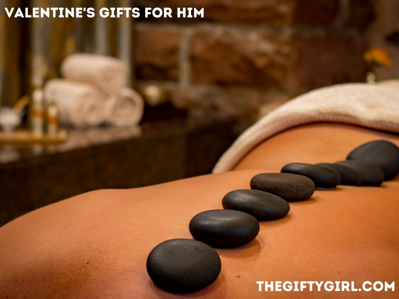 Image of a person with no shirt on lying on their stomach with hot stones in a line on their back for a hot stone massage. Text Overlay says, "Gift Ideas for Him, TheGiftyGirl.com"