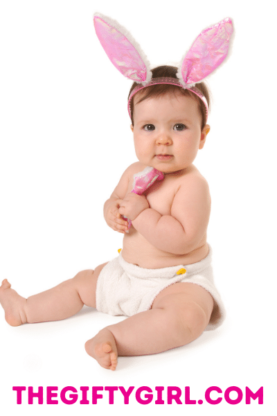 A baby wearing pink Easter Bunny Ear headband. Baby is wearing a white cloth diaper and looking at the camera. She is holding a pink toy and looking at the camera. 