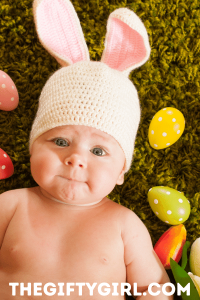 Baby wearing a crocheted bunny hat lying on green carpet scattered with colorful Easter eggs. The baby is looking up at the camera and chewing on his/her lip.
