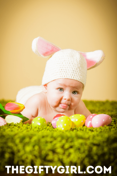 A young baby is lying on green carpet that has Easter eggs and tulips on it. Baby is wearing a crocheted bunny hat with ears and lying on his/her tummy looking at the camera.