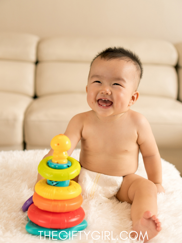 A baby boy in a diaper sits in front of a cream colored couch. He has a huge smile on his face and is sitting in front of a colorful stacking baby toy. 