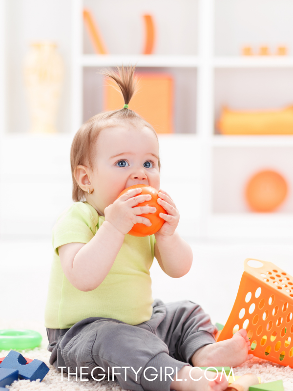 A baby girl with a ponytail on top of her head chews on a ball. She is sitting in front of an orange toy. 