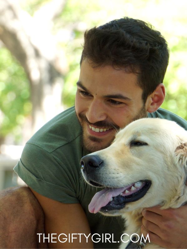 A man squats down next to a yellow Labrador dog. They are looking off camera and the man is smiling. 