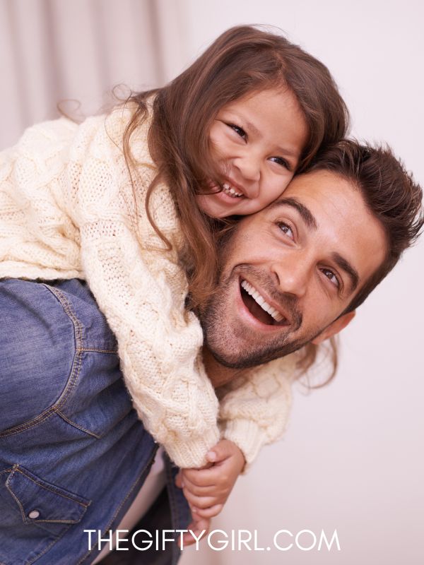 A dad holding a young girl on his back, piggyback style. He is looking up at her and laughing. 
