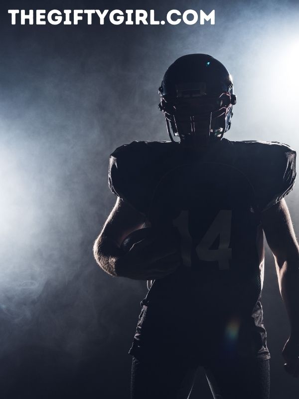A football player stands in shadows, only his silhouette showing. He is holding a ball and wearing a helmet. 