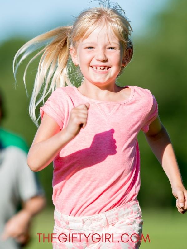A blond girl in a pink shirt is outside running toward the camera. She is smiling and her ponytail is blowing in the wind.