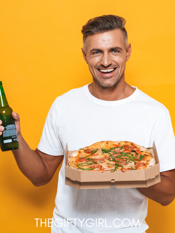 A man is holding a pizza in one hand and a drink in the other. Standing in front of an orange background, wearing a white shirt and smiling at the camera. 