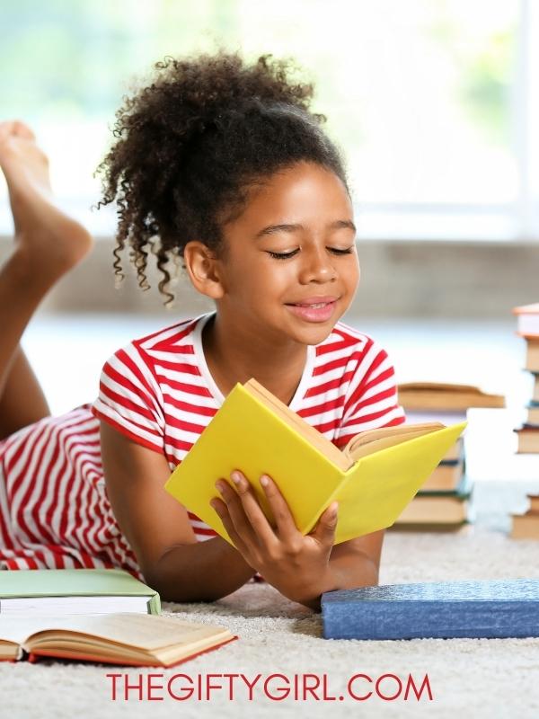 A black girl with red and white striped pajamas is lying on her stomach reading a yellow book. Lots of other books are stacked around her. She has a ponytail and is smiling. 