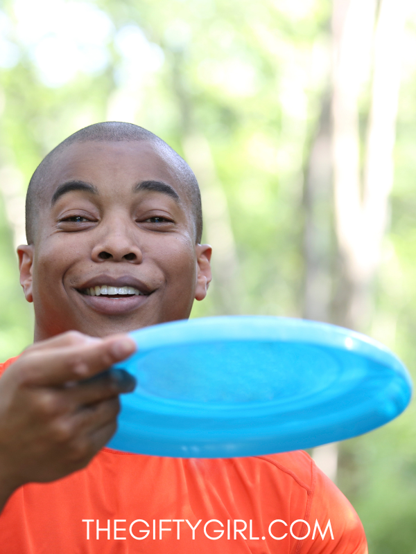 a black man wearing an orange shirt. He is outside with trees in the background. He is smiling at the camera and throwing a blue frisbee. 