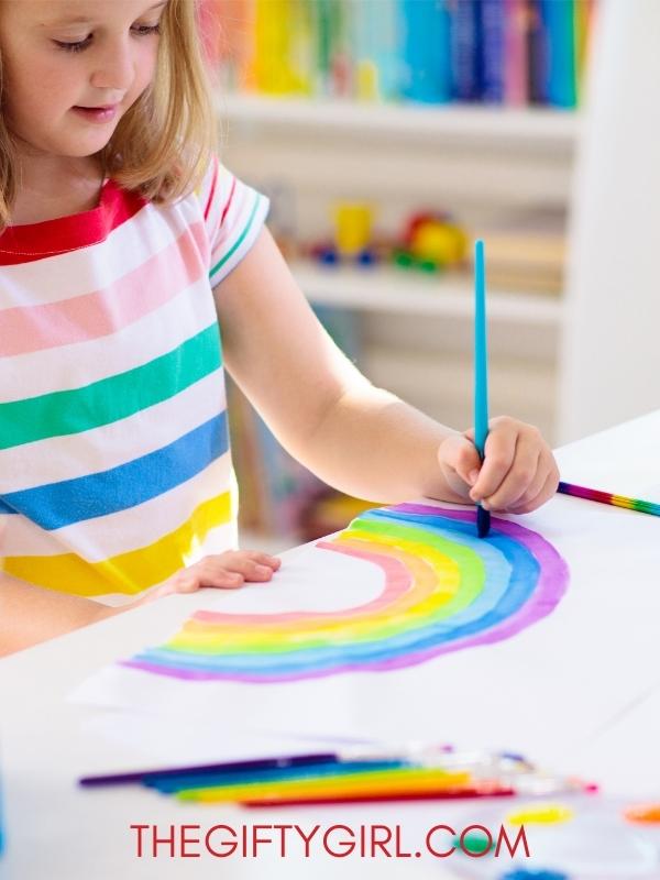 A blond girl wearing a rainbow shirt painting a rainbow on a big piece of paper on a white table. There is a bookshelf behind her with colorful books. 