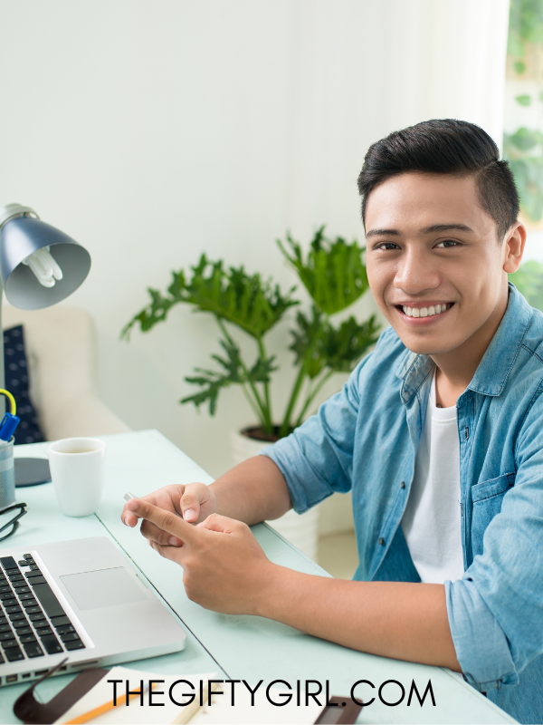 A younger man is sitting at a desk in front of a computer. He is smiling at the camera. There is a plant behind him.