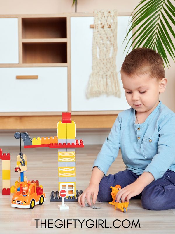 A 3-year-old boy sits on his knees in front of a car and lego building set. He is looking down at the toys. 