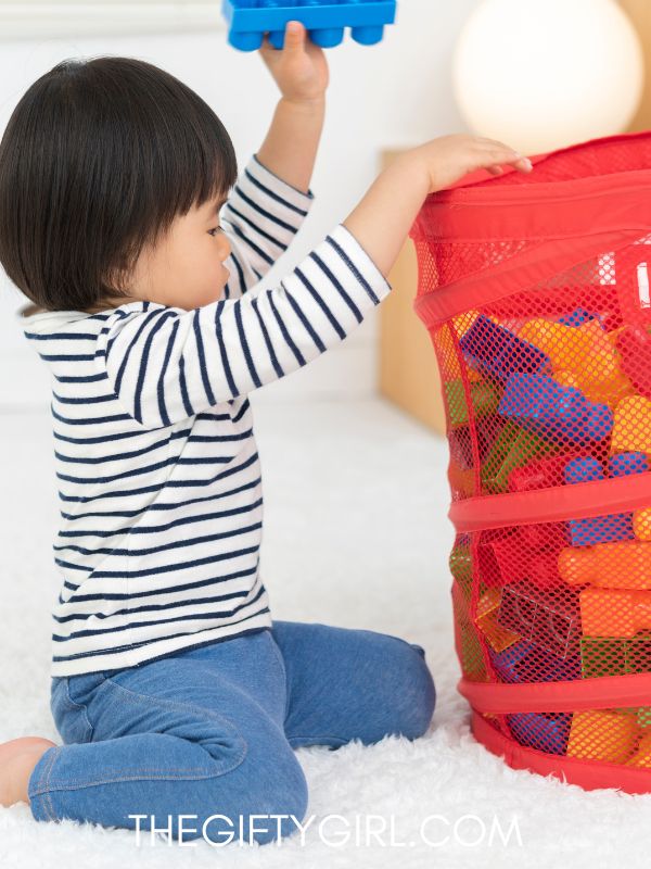 A little Asian girl kneels on a white rug. She is wearing a black and white striped shirt and is pulling building blocks out of a red mesh container.
