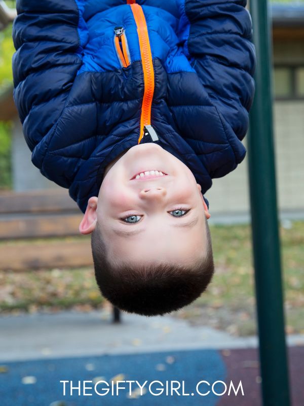 a 4-year-old boy hangs upside down from playground equipment. He is wearing a blue coat with an orange zipper and is smiling at the camera.