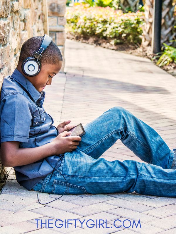 A black teen boy sits outside leaning against a wall. He is wearing headphones and looking down at a screen. 