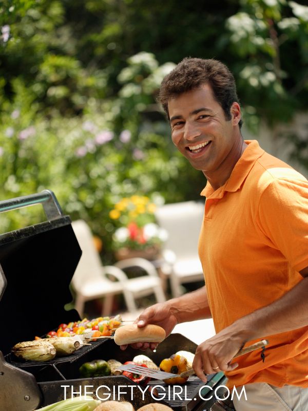 A man wearing an orange shirt grills outside. He is looking back at the camera and smiling.