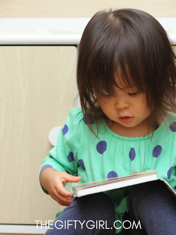 A two year old girl wearing a green polka dot shirt looks down at a book in her lap. Here Hair is dark and she has long bangs. 