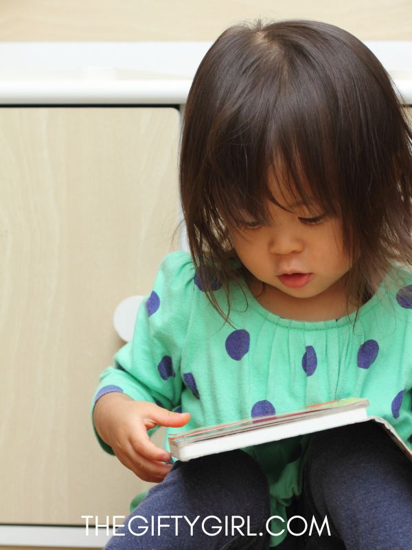 A two year old girl wearing a green polka dot shirt looks down at a book in her lap. Here Hair is dark and she has long bangs.