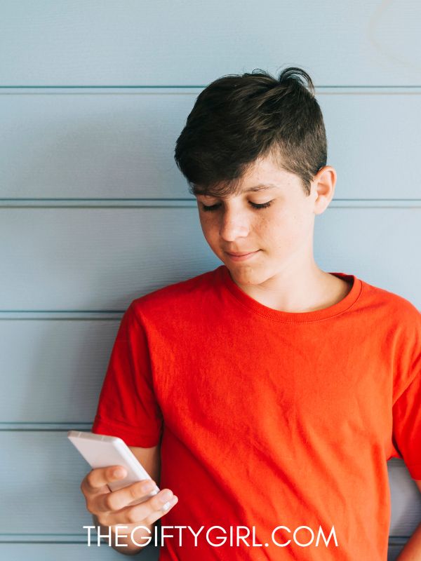 A teen boy in an orange shirt stands in front of a gray wall looking down at a phone. 