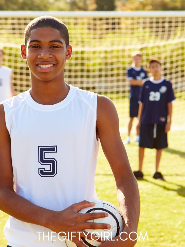 A teen boy with dark skin stands in front of a soccer net. he is wearing a white jersey and holds a soccer ball. he is smiling and looking at the camera. 