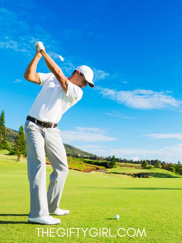 A man wearing a hat swings back a golf club on a gorgeous golf course. The grass is bright green, the sky is blue, it is a beautiful day.