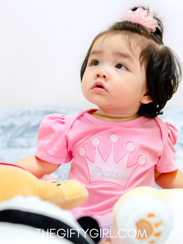 A baby girl wearing a pink princess shirt sits in a pile of stuffed animals. She is looking up at someone. Her hair is in a ponytail.